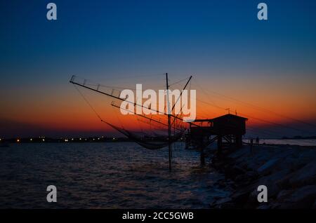 Traditionelles Fischerstationshaus mit Netz im Wasser der Adria am Pier Diga Sottomarina, Skyline mit Strand, erstaunlicher gelb-roter Sonnenuntergang in der Dämmerung, Dämmerung, Abendansicht, Norditalien Stockfoto