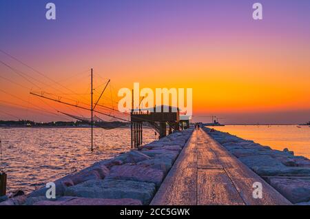 Traditionelles Fischerstationshaus mit Netz im Wasser der Adria am Pier Diga Sottomarina, Skyline mit Strand, erstaunlicher gelb-roter Sonnenuntergang in der Dämmerung, Dämmerung, Abendansicht, Norditalien Stockfoto