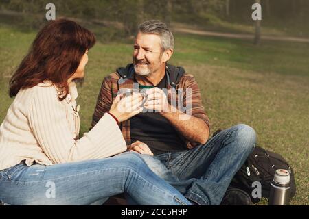 Glückliches Paar mittleren Alters verliebt auf einer Decke im Gras im Park sitzen, einander anschauen, lächeln und trinken Tee oder Kaffee aus Thermoskannen und Tassen. Stockfoto