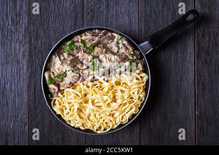 Rind Stroganoff mit Eiernudeln in einer Pfanne auf einem dunklen Holztisch, flach liegend, freier Platz Stockfoto