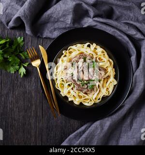 Rind Stroganoff gekocht mit saurer Sahne Pilze Sauce, serviert mit Tagliatelle in einer schwarzen Schüssel mit goldenen Gabel und Messer auf einem dunklen Holztisch, Klo Stockfoto