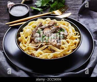 Rind Stroganoff gekocht mit saurer Sahne Pilze Sauce, serviert mit Eiernudeln in einer schwarzen Schüssel mit goldenen Gabel und Messer auf einem dunklen Holztisch, Clo Stockfoto