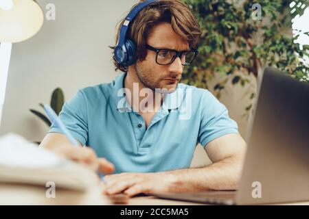 Junger Mann in blauem Hemd sitzt am Tisch und studiert mit Laptop Stockfoto