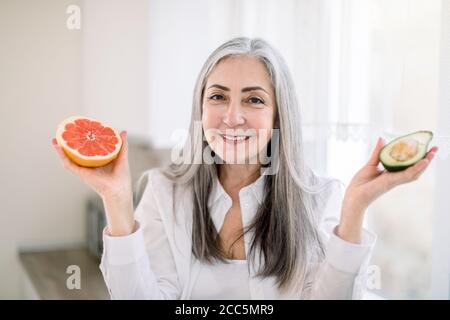 Gesunde Ernährung für Senioren, Obst-Food-Konzept. Ziemlich lächelnd ältere grauhaarige Frau sitzt auf der Arbeitsplatte in der modernen Küche, hält in den Händen Stockfoto