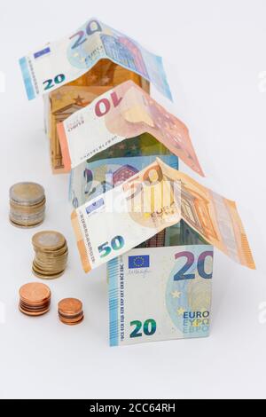 Häuser aus 10, 20 und 50 Euro-Banknoten, Stapel von Euro-Münzen, weißer Hintergrund. Konzepte von Preisen und Kosten im Zusammenhang mit Wohnen und Immobilien Stockfoto