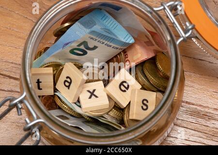 Euro-Münzen und Banknoten in einem Glas, mit dem Wort "Steuern" in Holzbuchstaben Stockfoto