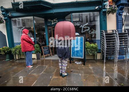 Tenby, West Wales. August 2020. UK Wetter: Tenby, West Wales. August 2020. Zwei Frauen Schlange im Regen vor einem Café auf der Hauptstraße in Tenby, West Wales an einem Sommertag stürmen und schlechtes Wetter Südengland und West Wales, Großbritannien. Mittwoch, 19. August 2020. Tenby, Pembrokeshire, West Wales, UK Kredit: Jeff Gilbert/Alamy Live News Stockfoto