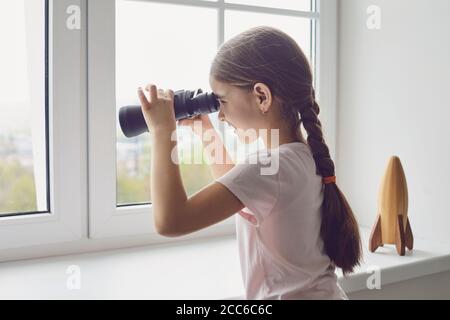 Home Isolation Konzept während Pandemie-Epidemie. Ein Mädchen mit Fernglas schaut aus dem Fenster, während es im Zimmer steht. Stockfoto