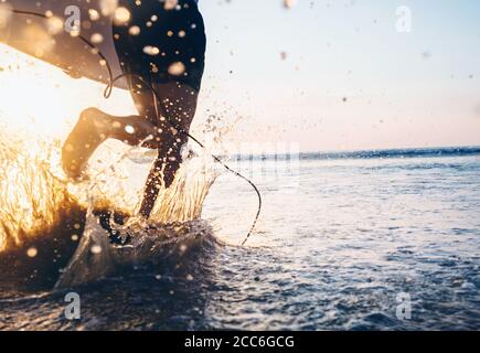 Man Surfer laufen im Meer mit Surfbrett. Nahaufnahme Wasserspritzer und Beine, Sonnenuntergang Licht Stockfoto