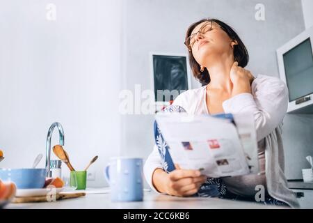 Eine Frau hat Nackenschmerzen, wenn sie morgens in der Küche ein Magazin liest Stockfoto