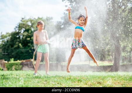 Heiße Sommertage Aktivität: Glückliches kleines Mädchen springt unter Wasser, wenn Bruder gießt sie aus Gartenschlauch Stockfoto