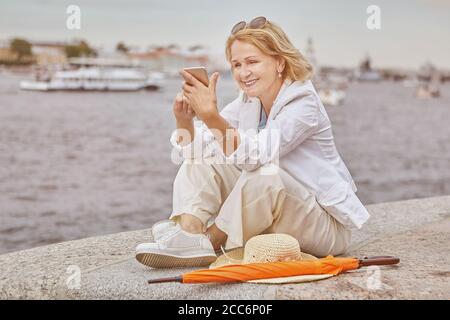 Schöne ältere kaukasische aktive Geschäftsfrau etwa 60 Jahre alt sitzt in der Nähe des Flusses mit Smartphone und lächelt. Im Alter hübsche Dame in lässig und Stockfoto