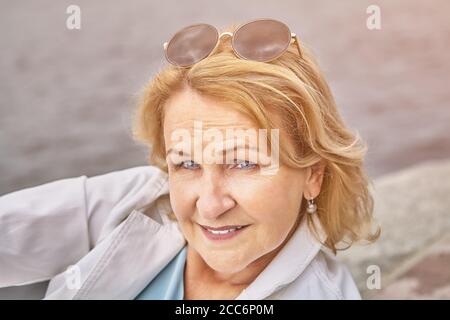 Attraktive Senior aktive kaukasische Frau über 62 Jahre alt mit blonden Haaren und Sonnenbrillen hat eine Pause im Freien in der Nähe des Flusses. Schöne ältere Leute, die freundlich sind Stockfoto