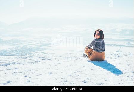 Glücklich lächelnde Frau sitzt auf dem verschneiten Berg Hügel über der Stadt. Sonniger Winter schönes Wetter Tag Stockfoto