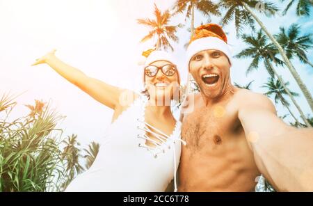 Weihnachten Bild von fröhlichen jungen Menschen paar gekleidet roten Santa Hüte machen Selfie mit tropischen Palmen Hintergrund. Stockfoto