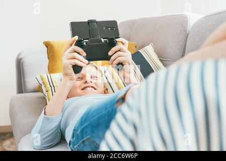 Teenager Junge liegt auf dem gemütlichen Sofa und spielt begeistert Spiel mit dem Gamepad mit Smartphone Stockfoto