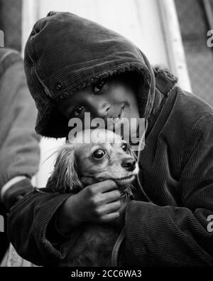 70ER JAHRE LÄCHELNDER AFROAMERIKANISCHER JUNGE IN CORDJACKE MIT KAPUZE UMARMT COCKER SPANIEL HUND BEIM BLICK AUF VAMERA - D6041 HAR001 HARS COCKER B&W AUGENKONTAKT GLÜCK SÄUGETIERE AFROAMERIKANER AFROAMERIKANER ECKZÄHNE COCKER SPANIEL CORD SCHWARZ ETHNIZITÄT POOCH FREUNDLICH PERSÖNLICHE BINDUNG ZUNEIGUNG HUND EMOTION JUVENILES SÄUGETIER SCHWARZ UND WEISS HAR001 ALTMODISCH AFRIKANISCH AMERIKANER Stockfoto