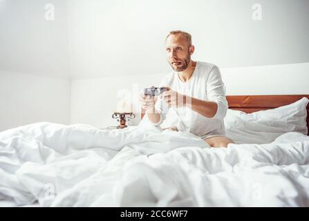 Großer kleiner Junge: Erwachsener Mann wachte auf und spielt im Bett sitzende PC-Spiele Stockfoto