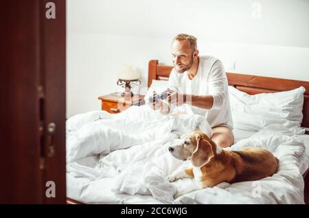 Mann spielt PC-Spiele sitzen im Bett am Morgen. Sein Beagle Hund beobachtet das Spiel mit sehr Interesse. Stockfoto
