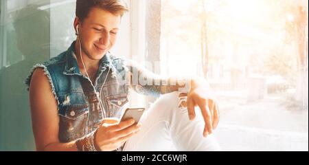 Junger Mann hört Musik mit Kopfhörern und Smartphone auf Fensterbank sitzen Stockfoto