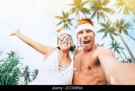 Fröhliche junge Leute paar gekleidet roten Santa Hüte machen Selfie Mit tropischen Palmen Hintergrund Weihnachtsbild Stockfoto