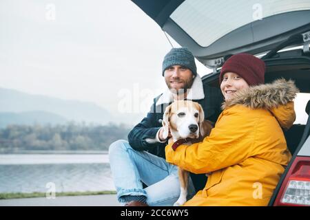 Vater und Sohn mit Beagle Hund sitzen zusammen im Kofferraum und lächeln in die Kamera. Stockfoto