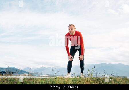 Der Weitwinkel-Shooting eines Mannes in rot gekleidet Langärmeliges Hemd sehr müde nach dem Joggen durch die Straße mit Berg Hintergrund und Ruhe zu lehnen forw Stockfoto