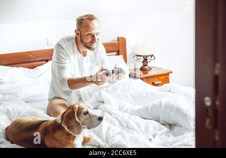 Erwachsener Mann wachte auf und spielt PC-Spiele im Bett sitzend, sein Hund beobachtet ihn mit großem Interesse Stockfoto