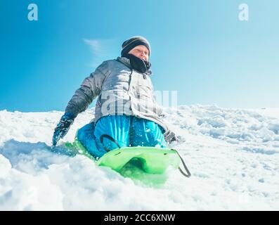 Happy Boy rutscht mit dem Schlitten vom Schneehügel herunter. Outdoor-Aktivitäten im Winter Stockfoto