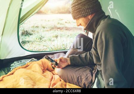 Bärtiger Reisender Warm Dressed man benutzt sein Handy im Zelt sitzend. Mobiltelefone mit Concept Image. Stockfoto