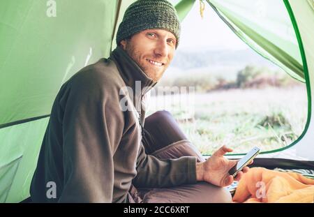 Bärtiger Reisender Warm Dressed man benutzt sein Handy im Zelt sitzend und lächelt in der Kamera. Mobiltelefone mit Concept Image. Stockfoto