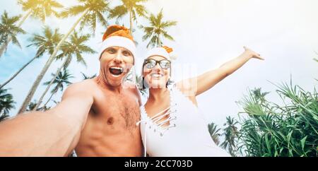 Weihnachten Bild von fröhlichen jungen Menschen paar gekleidet roten Santa Hüte machen Selfie mit tropischen Palmen Hintergrund. Stockfoto