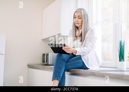 Moderne charmante Senior Dame mit langen grauen Haaren, trägt Jeans und weißes Hemd, sitzt auf der Küche Arbeitsplatte beim Surfen in sozialen Medien oder Stockfoto