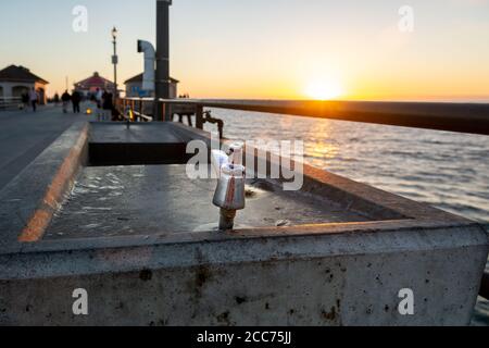 Sonnenuntergang über dem Pier und Meer mit Nahaufnahme von Edelstahl und Beton öffentlichen Trinkbrunnen auf dem Huntington Beach Pier. Stockfoto