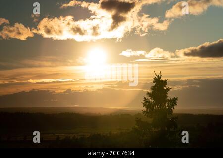 Luftaufnahme Sonnenuntergang über dunkelgrünem nordskandinavischen Wald - runde Streulicht, Wolken, Tannenspitze, warmer nebliger Abend, gelb-rote Sonne Stockfoto