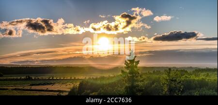 Luftpanorama des Sonnenuntergangs über hellgrünem nordskandinavischen Wald - runde Doppellinsenfackel, Wolken, Tannenspitze, warmer nebliger Abend, gelbe r Stockfoto