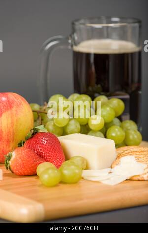 Gruyere Schweizer Käse, Becher Bier, Obst und Vollkorncracker auf einem hölzernen Schneidebrett Stockfoto