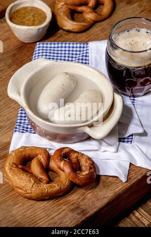 Münchner bayerische traditionelle Weißwürste in Keramikpfanne serviert mit deutschem süßen Senf, einem Becher dunklem Bier und Brezeln Brot auf weißer und blauer Serviette Stockfoto