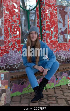 Eine Frau in blauem Denim Overall und einem grauen Hut Sitzt auf einer Bank in der Nähe einer Wand aus farbigem Glas Stockfoto