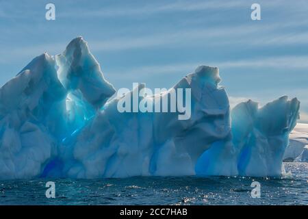 An einem sonnigen Nachmittag in der Antarktis schwebt ein Eisberg mit verschiedenen Blautönen im Meer nahe der gletscherbedeckten Küste. Stockfoto