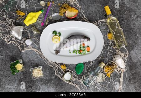 Weißer Fischteller mit Regenbogenforelle, Erbsen, Karotten, Zitronen und verschiedenen Abfällen im Fischernetz auf einem grauen Marmortisch Stockfoto