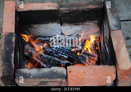 Schwelendes rickiges Feuerholz mit orangefarbenen Flammen zwischen Asche. Terrakotta-rote und rauchig schwarze verkohlte Ziegel in alten Kohlebrazier. Verkohltes Holz Stockfoto