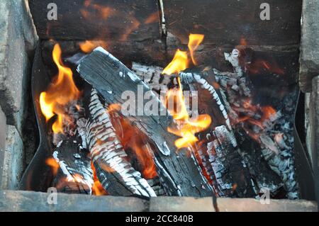 Schwelendes Feuerholz mit rissiger Oberfläche, bedeckt mit weißer Asche. Brennendes Holz Baumstämme Textur mit verschwommenen Flammen. Feuriger Hintergrund. Grillgrill concep Stockfoto
