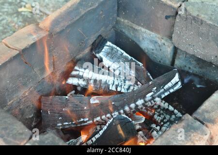 Schwelendes Feuerholz mit rissiger Oberfläche, bedeckt mit weißer Asche. Verkohlte Ziegel im alten mangal. Brennendes Holz Baumstämme Textur mit verschwommenen Flammen. Stockfoto