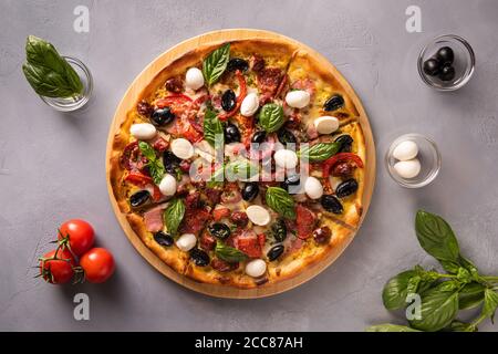 Italienische Pizza mit Schinken, Salami, schwarzen Oliven, Mozzarella-Käse, roten Tomaten und Basilikumblättern auf grauem strukturiertem Steinhintergrund und wenigen Zutaten i Stockfoto
