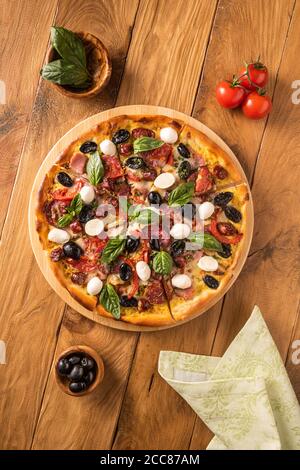 Italienische Pizza mit Schinken, Salami, schwarzen Oliven, Mozzarella-Käse, roten Tomaten und Basilikumblättern auf Holzgrund und wenigen Zutaten in Holzschüssel Stockfoto