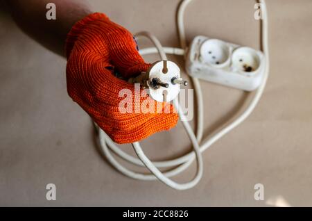 Die Hand trägt einen orangefarbenen Schutzhandschuh und hält nach dem Brand einen elektrischen Stecker. Weiße Steckdosenleiste nach Kurzschluss. Elektrische Sicherheit. Stockfoto