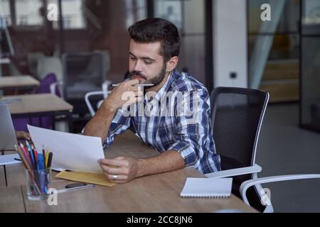 Junger seriöser kaukasischer Geschäftsmann oder Büroangestellter, der ein Dokument bei der Arbeit analysiert und an seinem Arbeitsplatz im modernen Coworking Space sitzt Stockfoto
