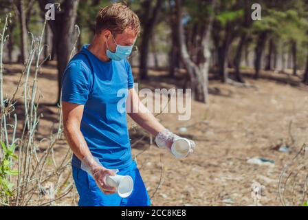 Volunteer in blauen Gesicht Maske Wald Sandstrand. Aktiver Mann abholen Müll Kunststoff Einweg Geschirr Taschen. Verschüttete Müll Müll Müll Planeten Verschmutzung Stockfoto