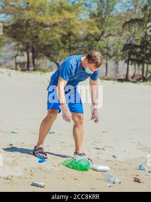 Volunteer in blauen Gesicht Maske Wald Sandstrand. Aktiver Mann abholen Müll Kunststoff Einweg Geschirr Taschen. Verschüttete Müll Müll Müll Planeten Verschmutzung Stockfoto